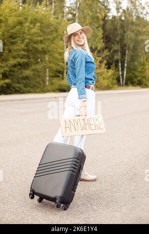 Junge Frau hält überall Karton mit Aufschrift, schwarzer Koffer in der Nähe des Waldes, geht entlang der Straße, schaut zurück. Stockfoto