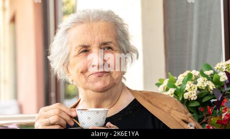 Alte lächelnde Frau, die türkischen Kaffee trinkt und sich auf dem Balkon entspannt. Ältere Frau im Jahr 70s lächelte und genoss die Zeit. Stockfoto
