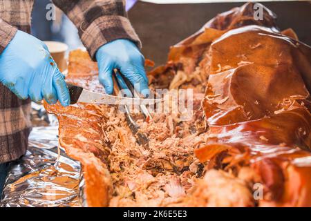 Nahaufnahme einer Person, die Fleisch mit Küchenutensilien schneidet und serviert, während sie blaue Handschuhe trägt Stockfoto