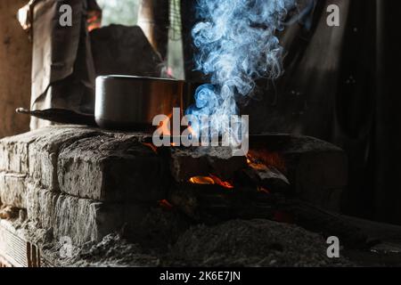 Nahaufnahme von brennendem Holz in einem Ofen aus Ziegel und grauer Asche. Blauer Rauch aus dem Holz, der als natürlicher Brennstoff zum Kochen von Speisen verwendet wird. Kochen Stockfoto