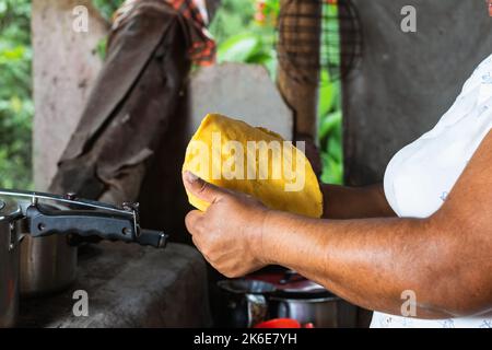 latina-Bäuerin, die in ihrer Küche eine traditionelle kolumbianische Arepa formte, um sie auf einem Backsteinofen zu grillen. Brünette Frau, die mit ihrer Hand einen Ara macht Stockfoto