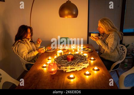 Symbolisches Bild Energiesparen, kalter Winter, Energiekrise, kalte Wohnung, Mutter und Tochter beim Abendessen warm angezogen, nur eine Lampe an, Kerzenlicht, Hitze Stockfoto