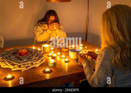 Symbolisches Bild Energiesparen, kalter Winter, Energiekrise, kalte Wohnung, Mutter und Tochter beim Abendessen warm angezogen, nur eine Lampe an, Kerzenlicht, Hitze Stockfoto