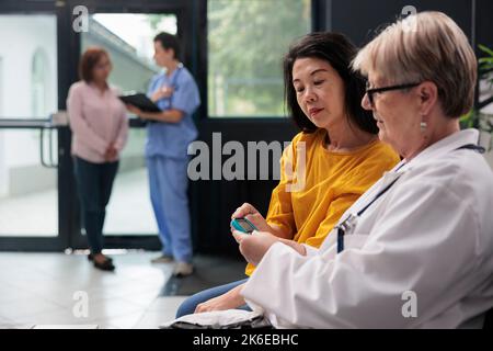 Multiethnische Frauen, die einen Insulinspiegel-Test mit Glucometer in der Wartehalle des Krankenhauses durchführen und eine Diabetesuntersuchung durchführen, um Glukose aus der Blutprobe zu messen. Arzt und alter Patient bei der Untersuchung. Stockfoto