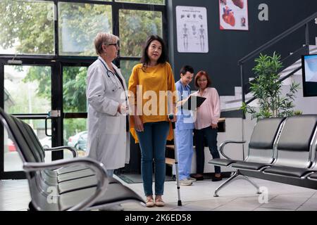 Leitender Arzt hilft verletzten Patienten, mit Stock zu gehen, leiden an körperlichen Beeinträchtigungen und mit Stock. Arzt, der an einer Rehabilitationstherapie teilnimmt, um sich von Beinverletzungen in der Einrichtung zu erholen. Stockfoto