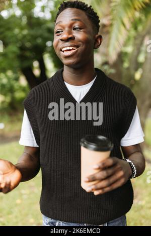 Junger afroamerikanischer Mann, der lächelt, um draußen auf der Straße eine Kaffeetasse zu trinken. Junge schwarze Studentin auf Kaffeepause im Park Stockfoto