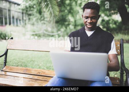 Junger schwarzer Mann in Weste, der im Freien an einem Laptop arbeitet, lächelnd auf den Bildschirm schaut. Student sitzt auf der Bank auf dem College-Campus, genießen freiberufliche Fernausbildung oder Fernausbildung Stockfoto