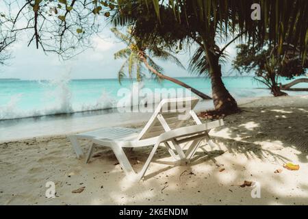 Blick auf ein Liegebett vor dem Meer in einem luxuriösen Resort auf den Malediven an einem warmen, sonnigen Tag umgeben von Palmen; ein Plastiksessel auf dem Korallenstrand Stockfoto