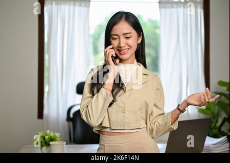 Elegante und charmante junge asiatische Geschäftsfrau oder Managerin ist in ihrem Büro und hat ein nettes Gespräch am Telefon mit ihrem Geschäftsparter. Stockfoto