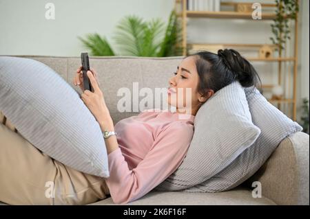 Entspannt und chillig verbringt die junge asiatische Frau ihr Wochenende zu Hause, legt auf der bequemen Couch und benutzt ihr Smartphone. Stockfoto