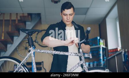 Starker gutaussehender Mann erfahrener Mechaniker konzentriert sich in seiner kleinen Werkstatt auf die Überprüfung und Reparatur des Lenkers eines gebrochenen Fahrrads. Qualifizierte Mitarbeiter und Wartungskonzept. Stockfoto