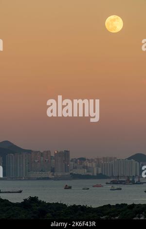 Der Vollmond steigt durch den orangefarbenen östlichen Himmel auf, wobei sich die untergehende Sonne in den Hochhäusern von Pol Fu Lam, Hong Kong Island, widerspiegelt Stockfoto