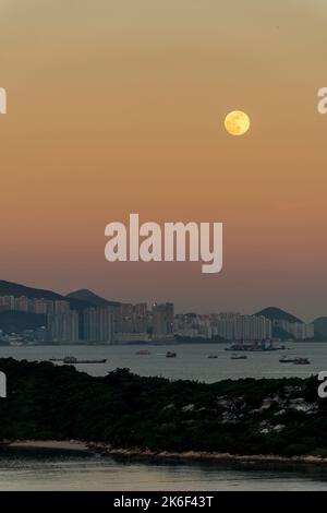 Der Vollmond steigt durch den orangefarbenen östlichen Himmel auf, wobei sich die untergehende Sonne in den Hochhäusern von Pol Fu Lam, Hong Kong Island, widerspiegelt Stockfoto