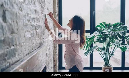Kreative junge Frau schmückt ihre Wohnung im Loft-Stil, indem sie Platz auf einer Ziegelwand für ein schönes Bild und eine Markierung mit Bleistift wählt. Kreativität und Raumkonzept. Stockfoto