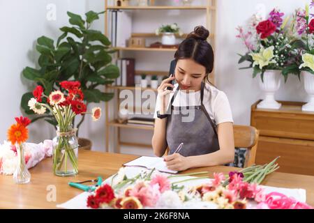 Floristen-Konzept, weibliche Floristin im Gespräch mit dem Kunden am Telefon und Notizen im Notebook im Geschäft. Stockfoto