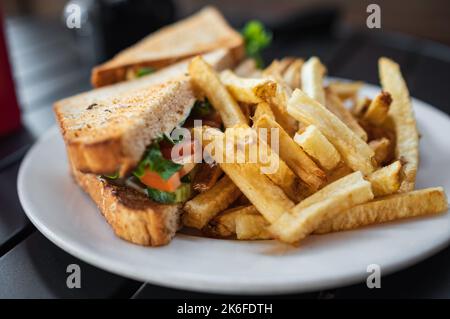 Club-Sandwich mit pommes auf weißem Teller. Gegrilltes Sandwich mit Gemüse, serviert mit Ketchup und gesalzenen Pommes frites. Niemand, verschwommener, selektiver Fokus Stockfoto