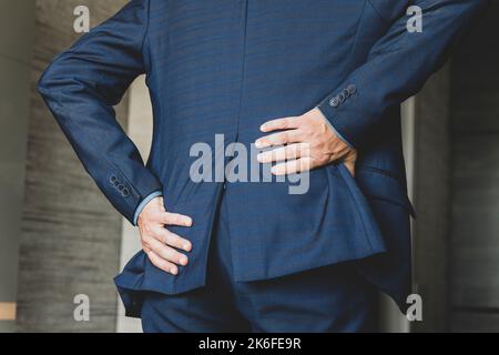 Geschäftsmann berührt seinen schmerzenden Rücken in seinem Büro. Büroangestellter hält seine Hand über seinen schmerzenden Rücken. Gesundheitliche Probleme durch sitzende Arbeit. Stockfoto