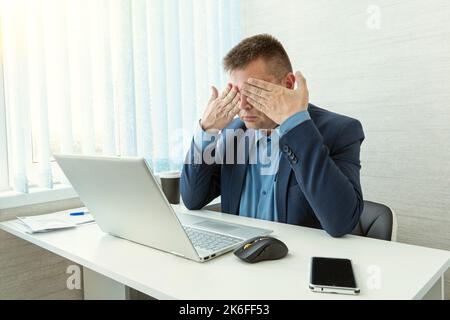Vorderansicht eines widerstrebigen jungen Mannes, der am Schreibtisch saß und das Gesicht mit der Hand bedeckte. Kerl mit geschlossenen Augen, der das Gesicht mit den Händen berührt, während er auf der Arbeit sitzt Stockfoto