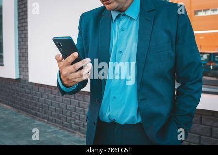 Executive Hand mit einem Smartphone auf der Straße mit einem Bürogebäude im Hintergrund. Ein Geschäftsmann in einem blauen Anzug sieht in seinem Smartphone ein Stockfoto