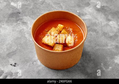 Kalte Gazpachosuppe mit Toast, getrocknete Kräuter in Papierschüssel auf grauer Steinoberfläche Stockfoto