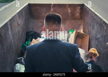 Ein verarmter Geschäftsmann rumpelt in einem Mülleimer auf der Suche nach Nahrung. Ein obdachloser Obdachloser in einem Anzug hält im Stehen einen Hamburger in der Hand Stockfoto