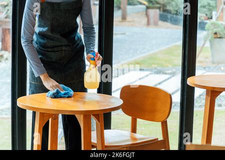 asiatische Frau Kellnerin Personal in Schürze Reinigungstisch mit Desinfektionsmittel Spray zum Schutz Infektion Coronavirus (covid-19) im Café Café Café Restaurant Stockfoto