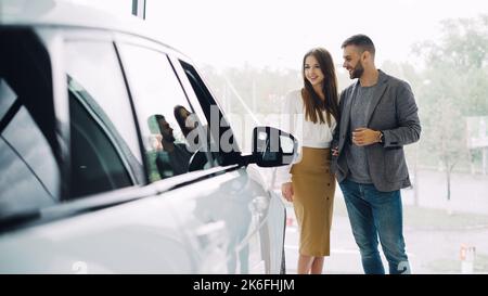 Attraktive junge Paar zuversichtlich bärtigen Kerl und seine lächelnde hübsche Freundin sind die Wahl neuer Automobil zusammen Blick auf Luxus-Auto in Autohaus und sprechen. Stockfoto
