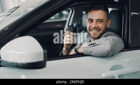Nahaufnahme eines hübschen bärtigen Typen von Neuwagenbesitzern, der in einem wunderschönen Auto sitzt und den Schlüsselanhänger hält und lächelnd die Kamera anschaut. Transport- und Personenkonzept. Stockfoto