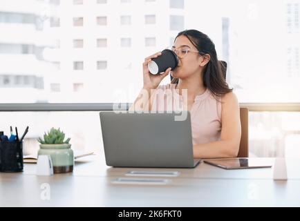 Ich brauche meinen täglichen Kraftstoff. Eine attraktive junge Geschäftsfrau, die allein im Büro sitzt und ihren Laptop benutzt, während sie Kaffee trinkt. Stockfoto