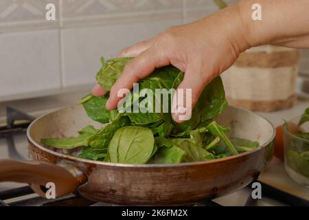 Nahaufnahme der Hand einer Frau, die Spinat in eine Bratpfanne wirft Stockfoto