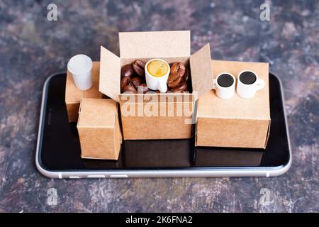 Mini-Versandkartons mit gerösteten Kaffeebohnen und Kaffeetassen, die auf einem Smartphone auf einem schmutzigen Hintergrund isoliert platziert werden. Kreative Kaffeeraue Stockfoto