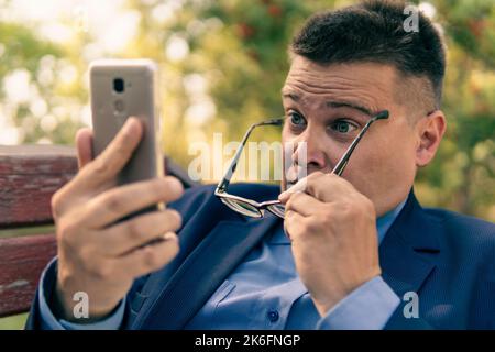 Besorgter älterer Mann, der eine Textnachricht am Telefon draußen im grünen Park liest. Schockierende Botschaft. Stockfoto