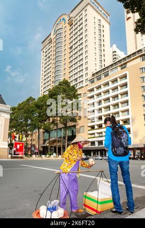 Vietnamesischer Straßenverkäufer mit Bambushut, der Kokosnüsse an Touristen verkauft, vor dem Caravelle Hotel, Ho Chi Minh City, Vietnam Stockfoto