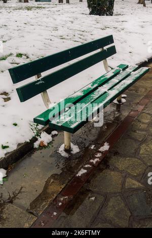 Tagesansicht einer leeren grünen Holzbank auf einem teilweise schneebedeckten Parkgelände. Stockfoto