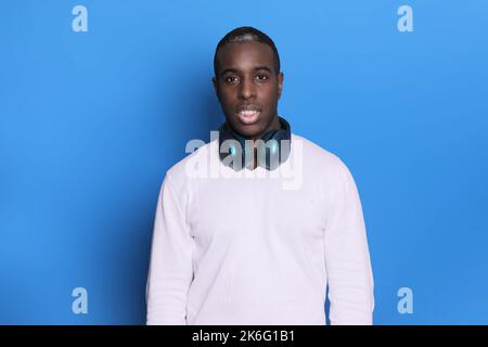 Foto eines gefühlvollen, dunkelhäutigen Teenagers, trägt blaue Kopfhörer um den Hals, schaut auf die Kamera, trägt einen warmen Winterpullover, isoliert auf blauem Rücken Stockfoto