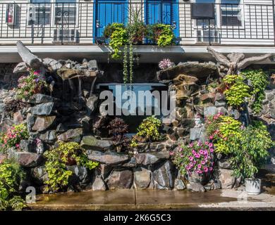 An einem Sommermorgen im Spring's Inn in Taylors Falls, Minnesota, USA, gibt es einen Steinbrunnen mit Blumen, Pflanzen, Steinvögeln und Elfenfiguren. Stockfoto