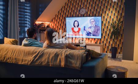 Paar sitzen auf dem Sofa im Wohnzimmer mit Snacks, schauen auf das Telefon, während TV-Nachrichten über die Ausstellung von neuen Smartphone, verbringen Wochenende zusammen in gemütlichen Hause. Live-TV-Übertragung. Stockfoto