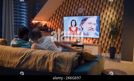 Paar sitzen auf dem Sofa im Wohnzimmer mit Snacks, schauen auf das Telefon, während TV-Nachrichten über die Ausstellung von neuen Smartphone, verbringen Wochenende zusammen in gemütlichen Hause. Live-TV-Übertragung. Stockfoto