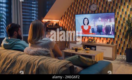Paar sitzen auf dem Sofa, reden, sehen Live-TV-Nachrichten über Cybersport, Cyberteam und Gaming-Meisterschaft. Frau und Mann ruhen sich am Wochenende zu Hause mit Pizza und Limonade aus. Live-TV-Übertragung. Esports. Stockfoto