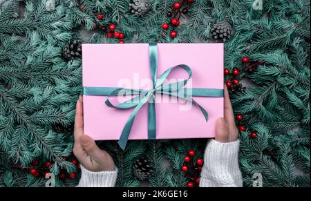 Frau Hände halten weihnachtsgeschenk oder Geschenkbox dekoriert Tannenbaum. Draufsicht auf die Hände einer jungen Frau, die ein Weihnachtsgeschenk in pi verpackt hält Stockfoto