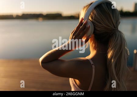 Rückansicht einer jungen, athletisch-blonden Frau in Sportkleidung, die am Sommermorgen kabellose Kopfhörer aufsetzt, um im Stadtpark zu laufen. Stockfoto