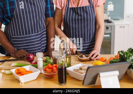 Älteres, vielseitiges Paar, das Schürzen trägt und in der Küche kocht Stockfoto