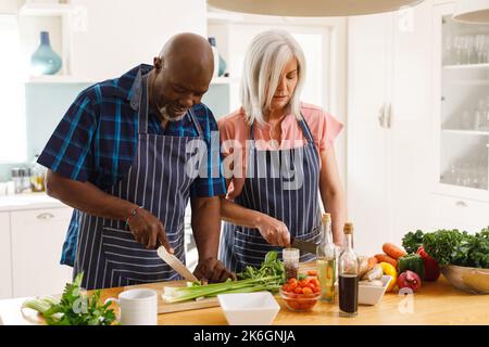 Glückliches, abwechslungsreiches Senior-Paar mit Schürzen und Kochen in der Küche Stockfoto