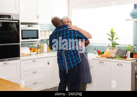 Glückliches, abwechslungsreiches Senior-Paar mit Schürzen und Tanz in der Küche Stockfoto