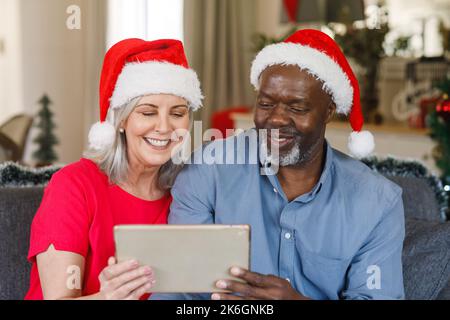 Glückliches, vielseitiges Senior-Paar mit weihnachtsmannmützen, die ein Tablet benutzen Stockfoto