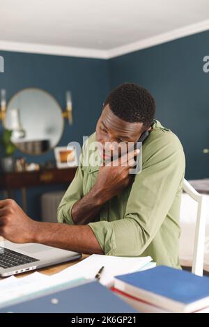 Vertikales Bild eines afroamerikanischen Mannes, der zu Hause im Schlafzimmer arbeitet, einen Laptop benutzt und am Telefon spricht Stockfoto
