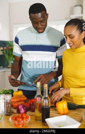 Vertikales Bild eines glücklichen, vielfältigen Paares, das das Rezept auf einem Tablet verwendet, um in der Küche zusammen Essen zuzubereiten Stockfoto