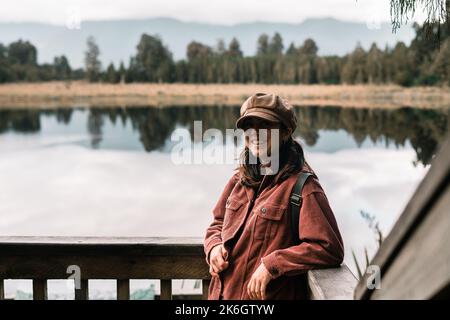 Blauäugige kaukasische Mädchen braun Hut Cord Jacke mit Taschen stehen glücklich lächelnd ruhig und entspannt lehnt auf Holzgeländer der Ruhe Stockfoto