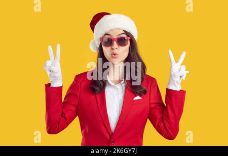 Überraschte Frau im Santa-Stil zeigt Siegesgeste mit zwei Händen auf gelbem Hintergrund. Stockfoto