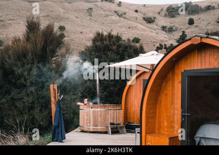 Kaukasischer Junge mit langen gesammelten Haaren sitzt in einem kleinen runden Holz-Whirlpool neben einer kleinen abgeschiedenen Holzhütte in der Mitte des Waldes in der Nähe Stockfoto